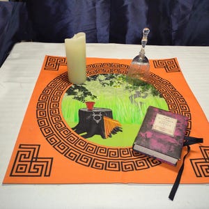 Puede incluir: Una alfombra naranja con un borde geométrico negro y una ilustración central de una escena de bosque, una vela, una campana y un libro. El libro tiene una cubierta morada y el título "Witchcraft & Spells".