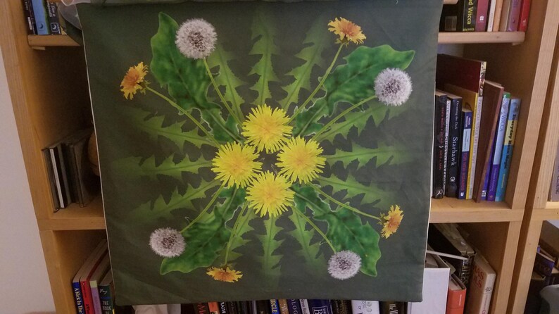 May include: A green canvas print with a symmetrical design of dandelion flowers and seed heads. The flowers are yellow and white, and the seed heads are white and fluffy. The background is a dark green.