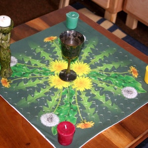 May include: A green table cloth with a dandelion pattern and a silver chalice in the center. There are four candles on the table cloth, two yellow, one green, and one blue.