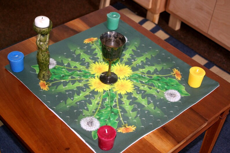 May include: A green table cloth with a dandelion pattern and a silver chalice in the center. There are four candles on the table cloth, two yellow, one green, and one blue.