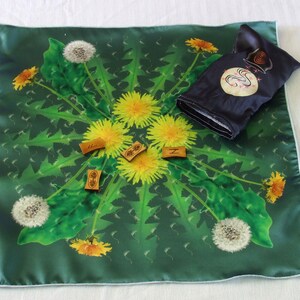 May include: A green fabric square with a dandelion design. The fabric has a white border. There are four small wooden tiles with letters on them. The tiles are arranged in a circle in the center of the fabric. There is a small black pouch with a white design on it.