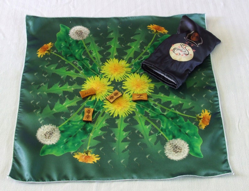 May include: A green fabric square with a dandelion design. The fabric has a white border. There are four small wooden tiles with letters on them. The tiles are arranged in a circle in the center of the fabric. There is a small black pouch with a white design on it.