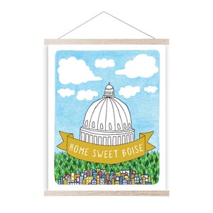 Puede incluir: Una impresión artística enmarcada con una ilustración dibujada a mano de un edificio con cúpula, probablemente un capitolio, con el texto "HOME SWEET BOISE" en una pancarta amarilla. El fondo incluye un cielo azul con nubes y un paisaje urbano.