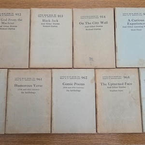 May include: A collection of ten vintage Little Blue Books, featuring titles like "The God From the Machine" and "Tales of the Strange and Supernatural." The books have off-white covers with black text, arranged on a wooden surface.