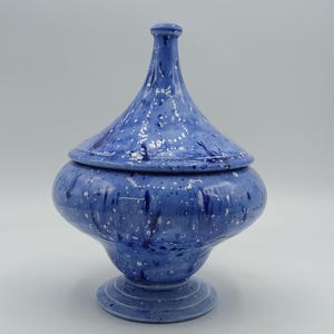 May include: A blue ceramic lidded jar with a speckled white and dark blue pattern. The jar features a rounded body, a pedestal base, and a tall, pointed lid with a small knob handle at the top, all finished in a glossy, mottled glaze.