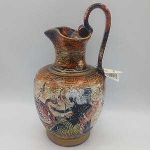 May include: A ceramic pitcher featuring a mottled brown and cream finish with a Greek key pattern around the neck. The body displays a black and white illustration of a figure seated beside a swan, with a tall handle and a small white tag attached.