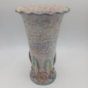 May include: A ceramic vase with a flared, scalloped rim and a textured, ribbed body in shades of cream, pink, and grey. The base is decorated with sculpted, three-dimensional floral and leaf motifs in muted tones of blue, green, and pink.
