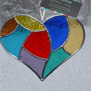 May include: A heart-shaped stained glass suncatcher featuring multi-colored textured glass segments in yellow, blue, red, orange, purple, teal, and amber, joined by silver metal solder lines and suspended by a white ribbon with a business card.