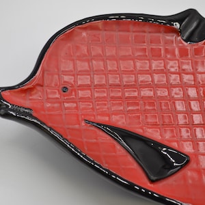 May include: A ceramic dish shaped like a fish with a glossy red interior featuring a raised grid pattern and a smooth black border. The dish includes a small black eye detail and a prominent black fin accent on the side of the red textured body.