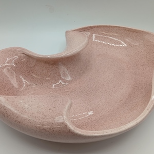 May include: A pale pink ceramic serving dish with a speckled finish and a unique, asymmetrical, curved shape. The bowl features deep, flowing indentations that create distinct sections within the smooth, glossy, glazed surface.