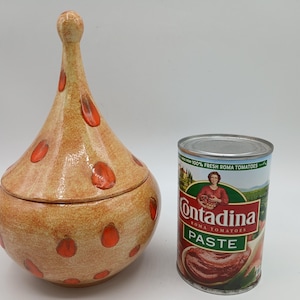 May include: A tan ceramic lidded jar with a pointed finial and orange teardrop-shaped spots, positioned next to a metal can of Contadina Roma Tomato Paste for scale. The jar has a rounded base and a matching lid with a tall, tapered top.