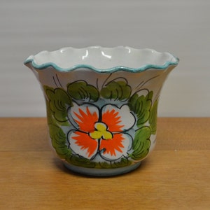 May include: A white ceramic flower pot with a scalloped rim and a turquoise trim. The pot is decorated with a large orange and yellow flower surrounded by green leaves. The pot sits on a wooden surface.