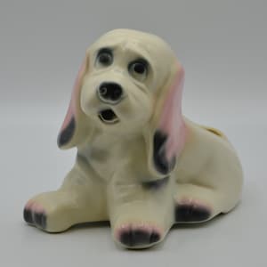 May include: A ceramic dog figurine in a seated position. The dog is primarily off-white with pink and black accents on the ears, paws, and nose. The dog has large, expressive eyes and an open mouth, giving it a surprised look.