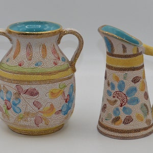 May include: Two ceramic vases with a crackle finish. One is a round vase with two handles, and the other is a pitcher. Both have a turquoise interior and are decorated with colorful floral patterns and gold accents. The vases are on a white background.