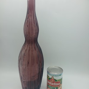 May include: A tall, purple glass bottle with a ribbed, vertical texture and a narrow neck, positioned next to a standard-sized metal can of Contadina tomato paste, which serves as a visual reference for the scale of the glass bottle.