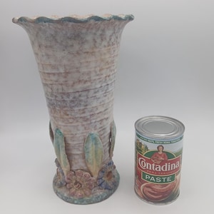 May include: A tall, textured ceramic vase with a flared top and floral base. The vase has a light brown and gray color scheme with blue and pink accents. A can of Contadina tomato paste is next to the vase.