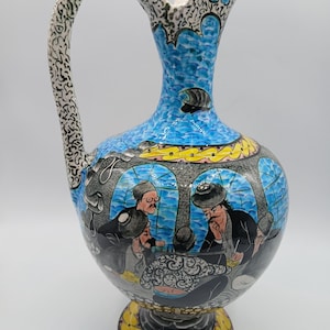 May include: A ceramic pitcher featuring a hand-painted scene of four figures seated around a table. The vessel has a blue upper section, a black base, and a handle decorated with intricate black and white patterns and yellow accents.