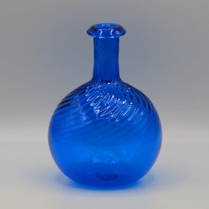 May include: A vibrant cobalt blue glass decanter featuring a spherical body and a slender, elongated neck. The glass surface displays a distinct, swirling ribbed texture that wraps around the entire vessel, ending in a smooth, circular rim.