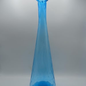May include: A tall, slender, translucent blue glass vase with a conical shape that tapers toward a narrow neck and a flared rim. The surface of the glass features a subtle vertical ribbed texture that runs from the base up to the neck.
