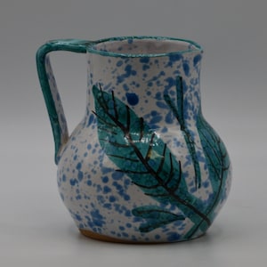 May include: A ceramic pitcher with a white base, speckled with blue paint. The pitcher features a teal handle and leaf design. The leaves are teal with black veins. The pitcher is a decorative item.