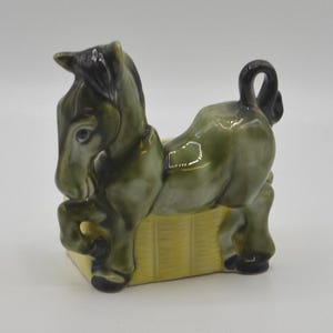 May include: A ceramic figurine of a dark green donkey with black hooves, mane, and tail, standing over a rectangular yellow base with a textured, grid-like pattern. The donkey is depicted in a prancing pose with one front leg raised.