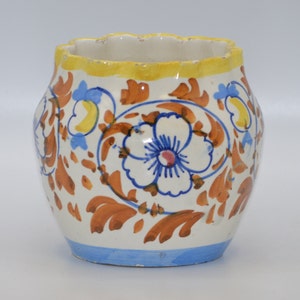 May include: A white ceramic vase with a scalloped yellow rim and a blue base. The vase is decorated with a floral pattern in orange, blue, and yellow. The design features a large white flower with blue and pink details.