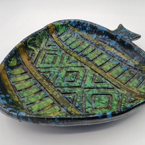 May include: A ceramic dish shaped like a pomegranate with a glossy finish. The surface features a textured geometric pattern in shades of green, blue, and black, with a speckled glaze effect throughout the interior and along the outer edges.