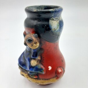 May include: A small ceramic vase featuring a glossy glaze in shades of black, red, blue, and yellow. The exterior surface is decorated with a raised, three-dimensional figure of a person in a blue outfit, integrated into the curved body of the vessel.