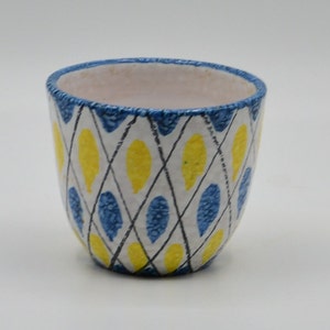 May include: A small, ceramic cup with a white interior and a blue rim. The exterior features a white background with a black diamond pattern, filled with yellow and blue teardrop shapes. The cup is approximately 3 inches tall.