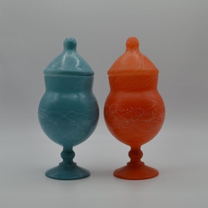 May include: Two decorative glass jars with lids. One is teal, the other is orange. Both have white splatter paint designs and are on pedestals. The jars are likely used for storage or display.