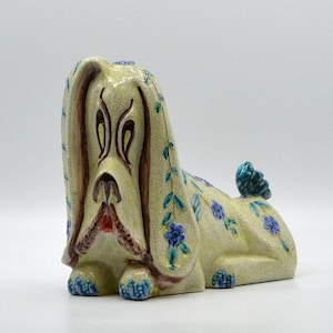 May include: A ceramic dog figurine with a cream-colored crackle glaze finish. The dog features long, drooping ears, brown painted facial details, and a red mouth. The body is decorated with small blue floral patterns and green leaves.