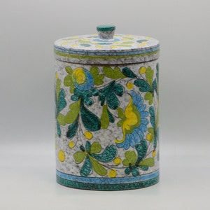 May include: A cylindrical ceramic container with a lid, featuring a floral design in shades of blue, green, and yellow against a speckled gray background. The container has a matching lid with a small knob and a blue border.