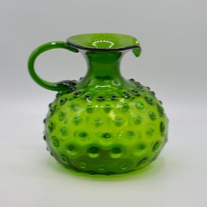 May include: A vibrant green glass pitcher with a rounded body and a handle. The pitcher has a textured surface with small, raised bumps. The rim is slightly flared and has a dark trim. This decorative pitcher is a unique piece of glassware.