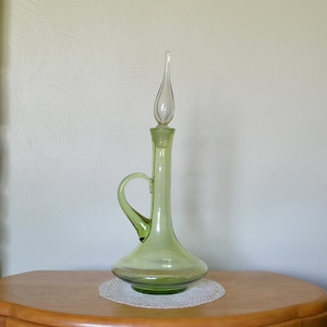 May include: A tall, light green glass decanter with a handle and a clear, teardrop-shaped stopper. The decanter sits on a white, circular doily. The design features a bulbous base and a slender neck.