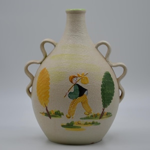 May include: Cream-colored ceramic vase with a textured surface, featuring a painted scene of a person carrying a bundle on a stick, with yellow and green trees. The vase has two sets of curved handles.