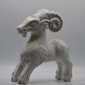 May include: A white ceramic figurine of a ram standing in a forward-leaning pose. The sculpture features detailed carved textures representing the animal's fur, a small beard, and large, prominent curled horns on the side of its head.