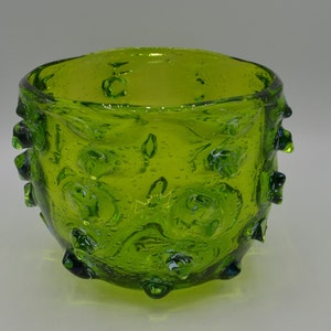 May include: A round, lime green glass bowl with a textured surface and bubble details. The bowl has a thick, irregular rim and small, rounded protrusions around the exterior. The glass appears translucent, with a lighter green interior.