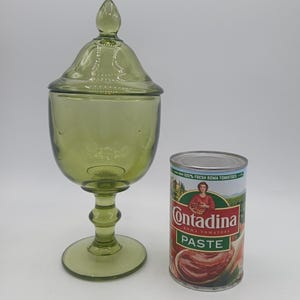 May include: A green glass apothecary jar with a lid, next to a can of Contadina Roma Tomatoes paste. The jar has a bulbous body, a stem, and a rounded base. The can is cylindrical with a red and green label.