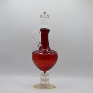 May include: A ruby red glass decanter with a clear glass stopper and handle. The decanter has a bulbous body and sits on a clear and white pedestal base. The handle is curved and attached to the neck of the decanter.