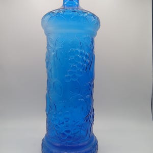 May include: A tall, vibrant blue glass bottle with a detailed grape and leaf pattern. The bottle has a rounded base, a cylindrical body, and a narrow neck. The glass has a frosted appearance, enhancing the intricate design.