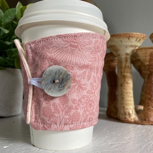 May include: A pink floral fabric coffee cup sleeve with a large, round, light-coloured button closure. The sleeve is wrapped around a white disposable coffee cup with a white lid. The background includes a green plant and ceramic decor.
