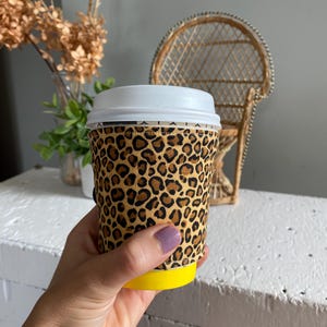 May include: A reusable coffee cup sleeve with a leopard print design. The sleeve is made of fabric and has a yellow bottom.