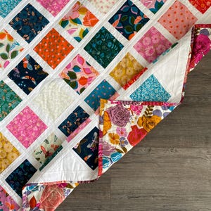 Peut inclure: Une courtepointe blanche avec un motif en losange. La courtepointe est faite de différents tissus colorés avec des motifs floraux et géométriques. La courtepointe est pliée en deux et le coin est relevé, révélant le dos de la courtepointe, qui est un tissu blanc avec un motif floral.