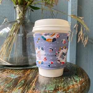 May include: A white coffee cup with a blue and white fabric sleeve featuring a pattern of small animals and flowers. The sleeve has a white polka dot background.
