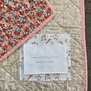 May include: A patchwork quilt featuring floral and dotted patterns in cream, pink, and gold. The quilt has a pink border and a white label with the text "Charlotte Elise Rowan April 21, 2017".