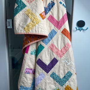 Peut inclure: Une courtepointe faite à la main avec un fond crème et des motifs en chevrons colorés. La courtepointe présente divers tissus dans les tons de rose, bleu, vert, jaune et orange. Les bords sont finis avec une bordure rose.