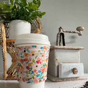 May include: A white coffee cup with a floral print fabric sleeve. The sleeve features a variety of pink, orange, yellow, and blue flowers on a white background. The cup is resting on a white surface with coffee beans scattered around it.