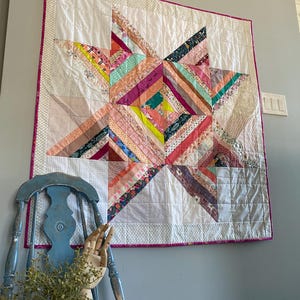 May include: A handmade quilt featuring a star design. The star is made from fabric strips in pinks, purples, oranges, and greens. It has a white background and a pink border. The quilt is displayed on a grey wall.