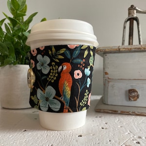May include: A white coffee cup with a black fabric sleeve featuring a colourful parrot and floral pattern. The sleeve has a gold button.