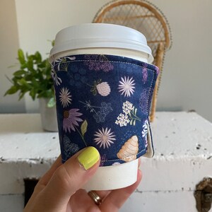May include: A white takeaway coffee cup with a navy blue fabric sleeve featuring a floral pattern in shades of pink, white, and green. The sleeve has a seam down the side.